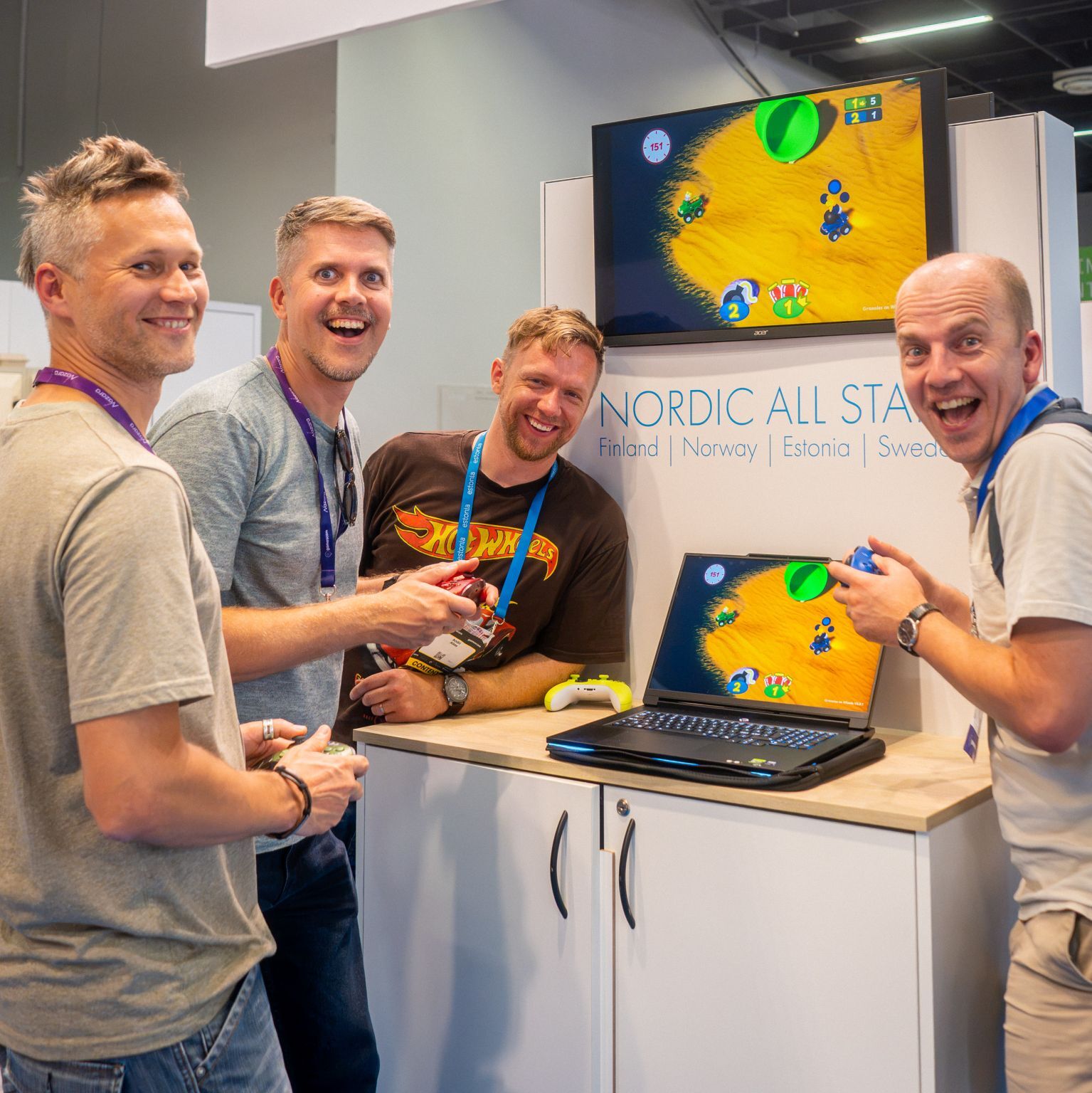 Neli naeratavat meest mängupultidega Gamescomi messil Nordic All Starsi boksis, taustal ekraanidel värviline autovõidusõidumäng.